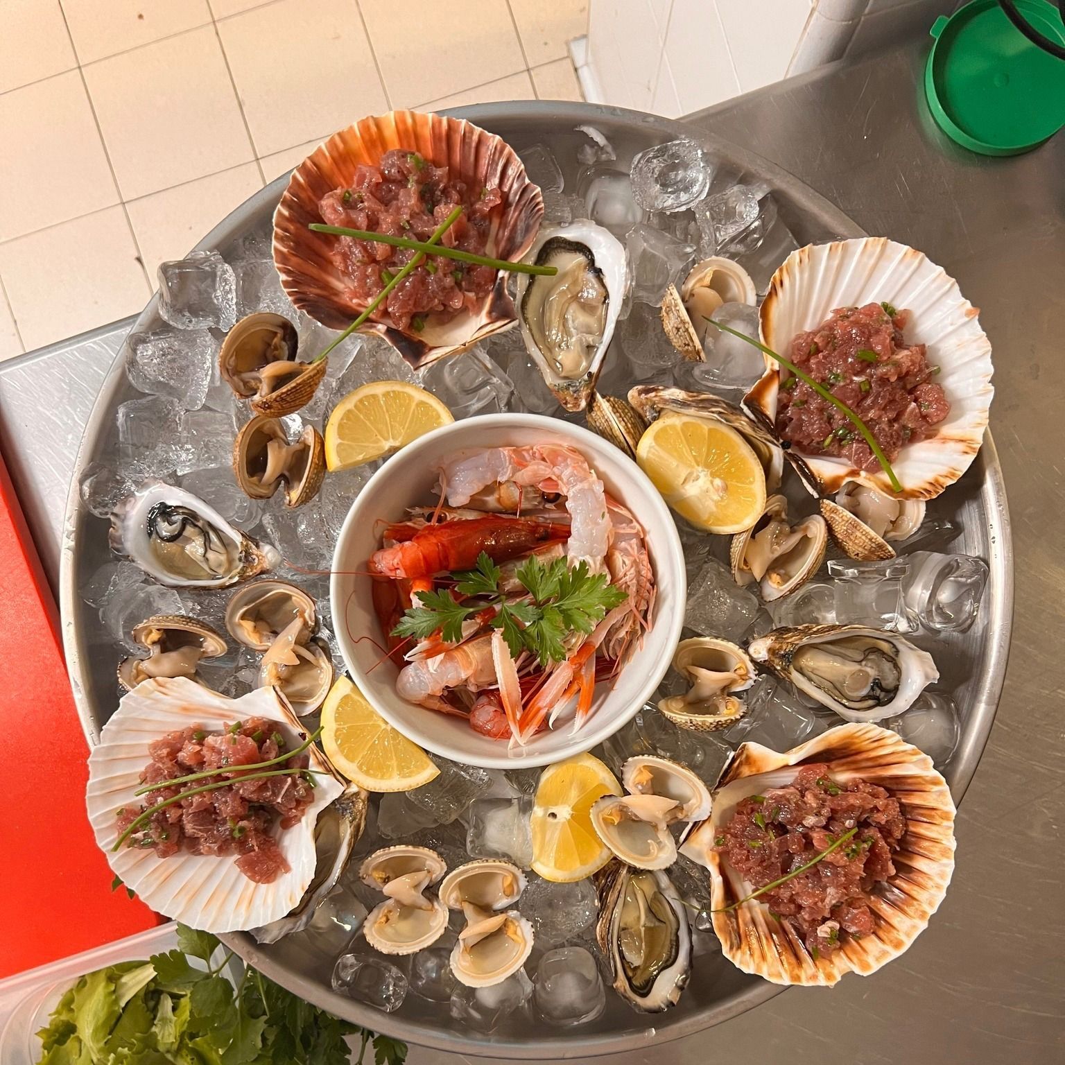 Piatto di frutti di mare: capesante crude, gamberi, ostriche, vongole, limoni e ghiaccio su un vassoio di metallo.