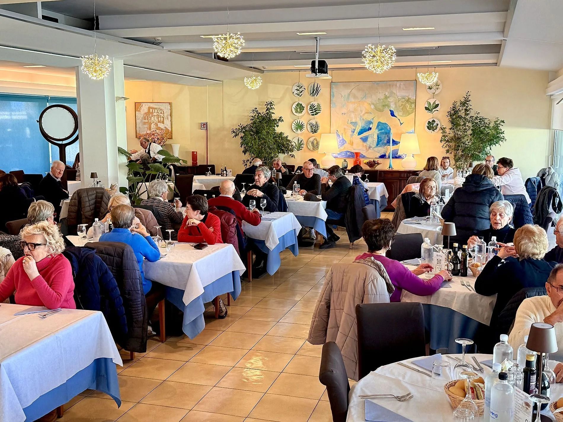 Interno di un ristorante, tavoli con persone che cenano. Tovaglie bianche, pareti dai toni neutri, luci decorative e finestre.