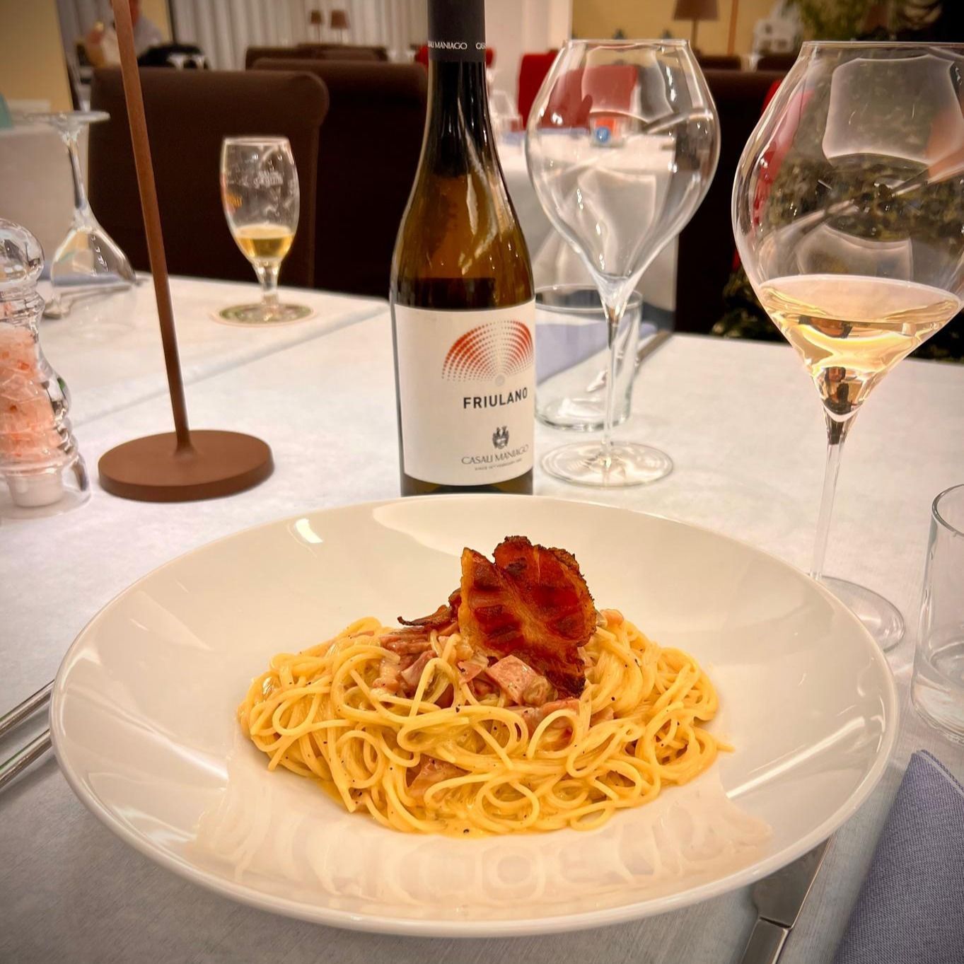 Spaghetti alla carbonara con pancetta su un piatto bianco, bottiglia di vino, bicchieri e un tavolo apparecchiato al ristorante.