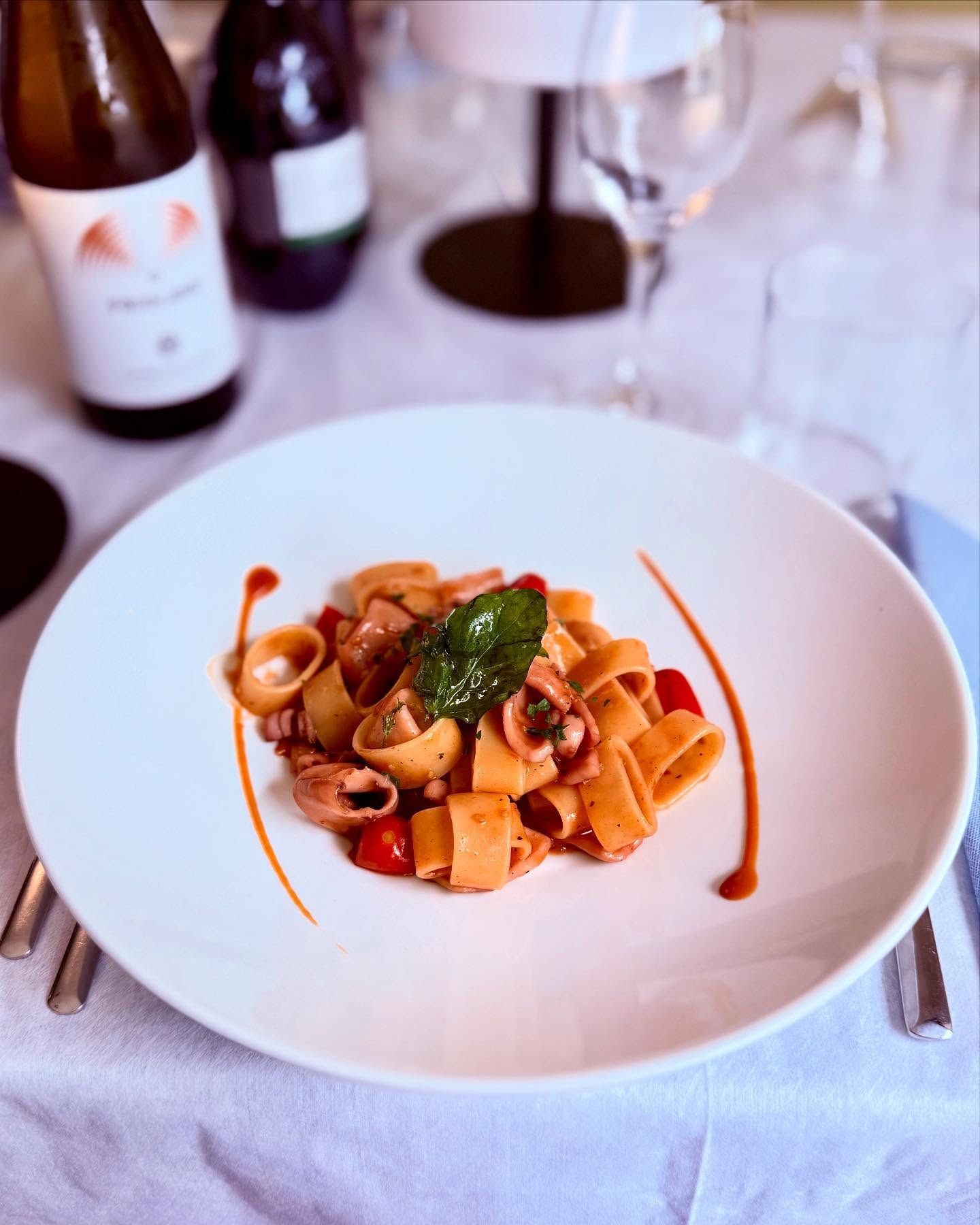 Piatto di pasta su un piatto bianco, con salsa di pomodoro, pomodori e basilico. Bottiglie di vino sullo sfondo.