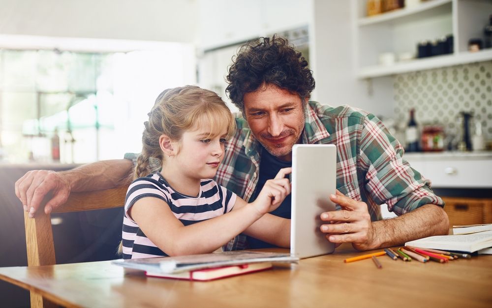 A man and a little girl are sitting at a table looking at a tablet.