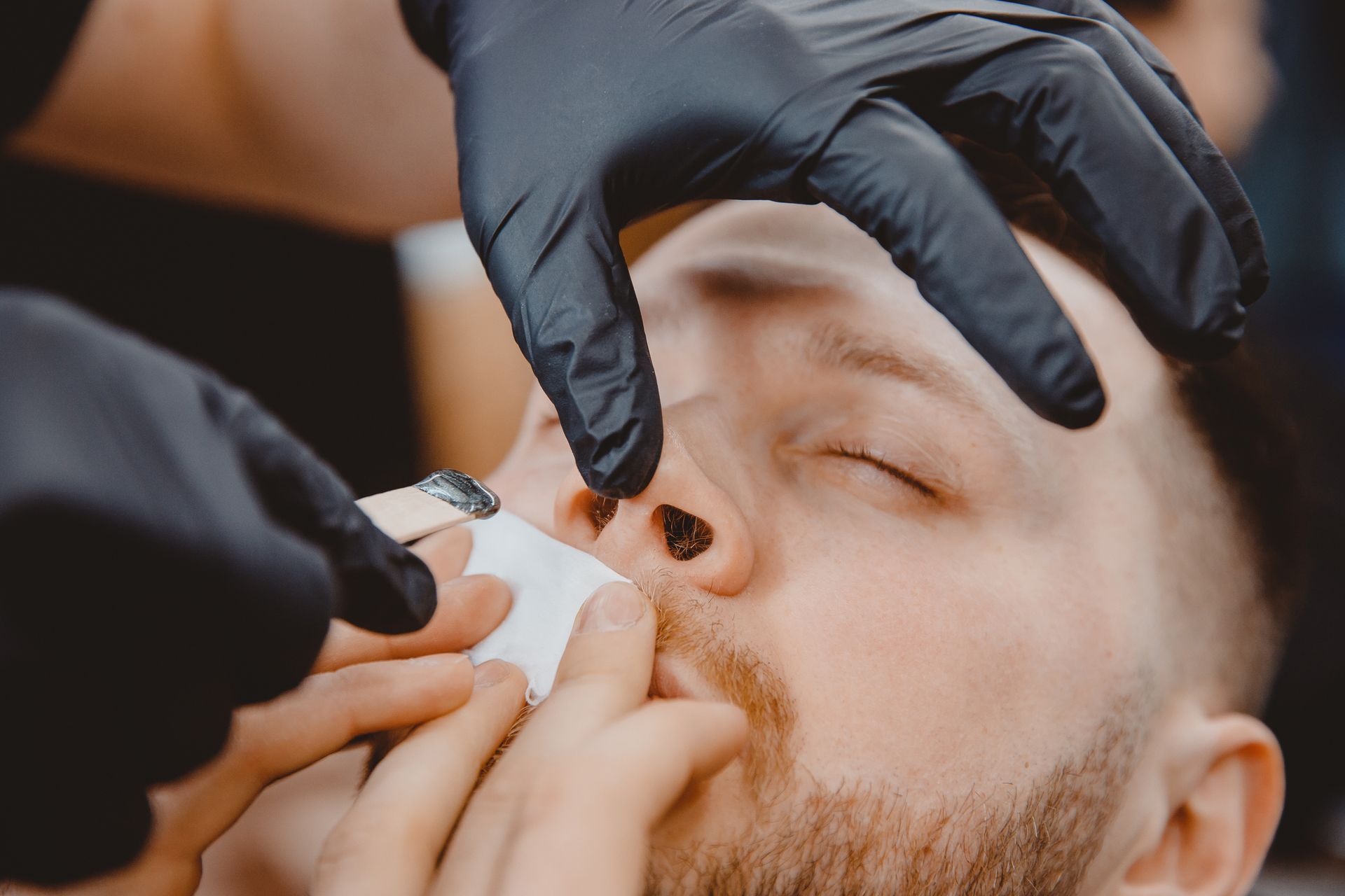 A man is getting his nose waxed by a barber wearing black gloves.
