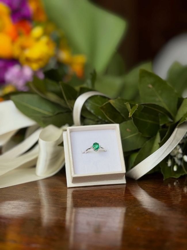 Un anello con smeraldo è custodito in una scatola bianca aperta, su una superficie di legno lucido, tra piante e nastri bianchi.