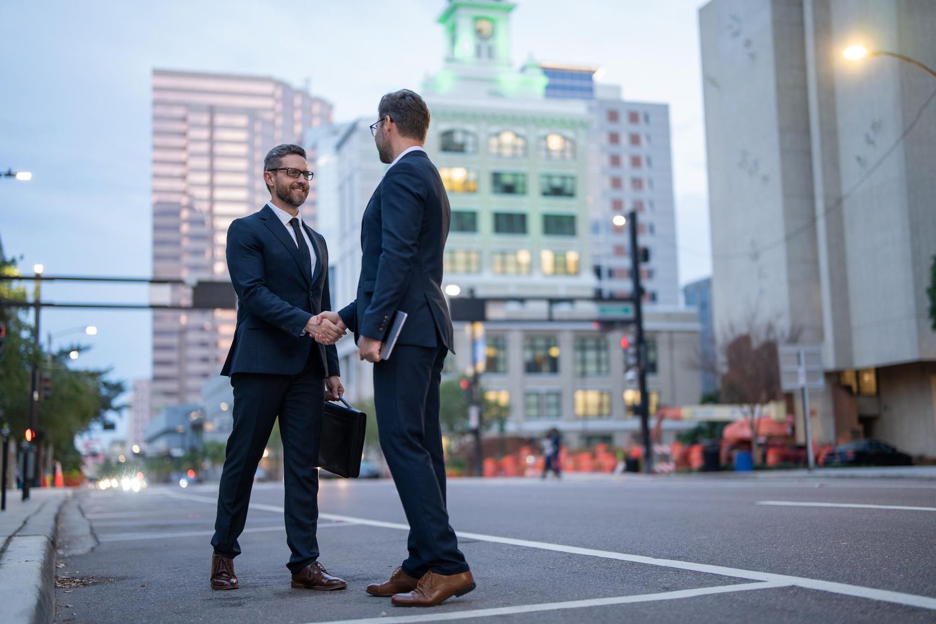 Two professionals in dark business suits shake hands on a city street at dusk, with office buildings in the background.