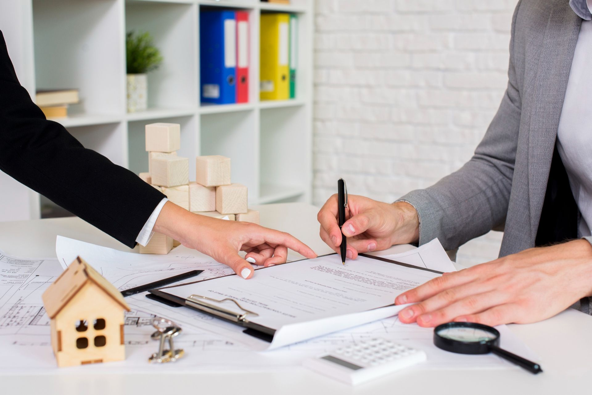 Hands gesture over a document on a desk with a small wooden house model, keys, a calculator, and a magnifying glass.