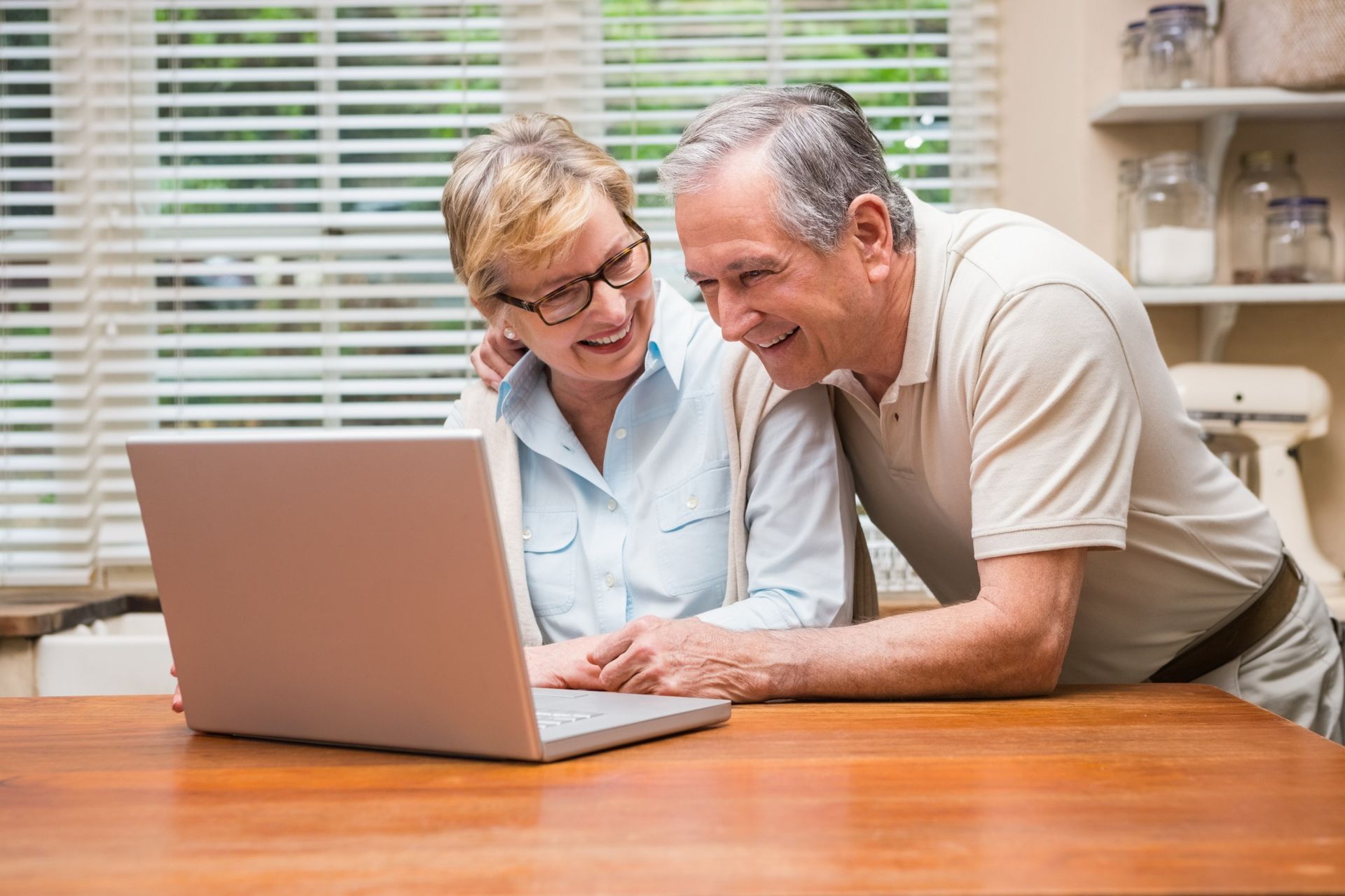 Two people sitting at a wooden kitchen table, smiling while looking together at a laptop screen.