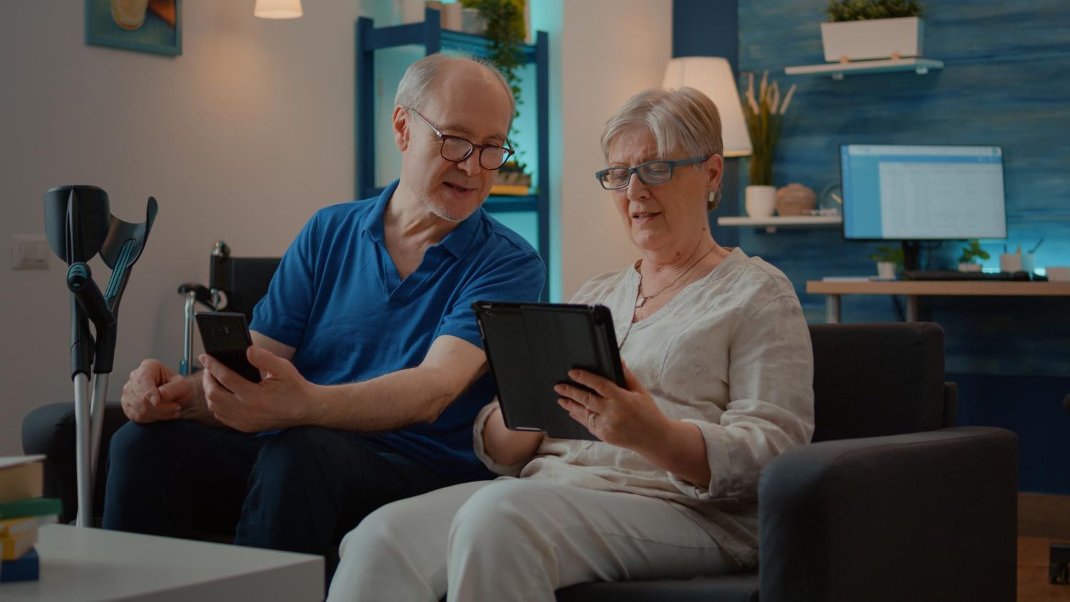 An older couple on a couch looking at a tablet and smartphone in a living room.