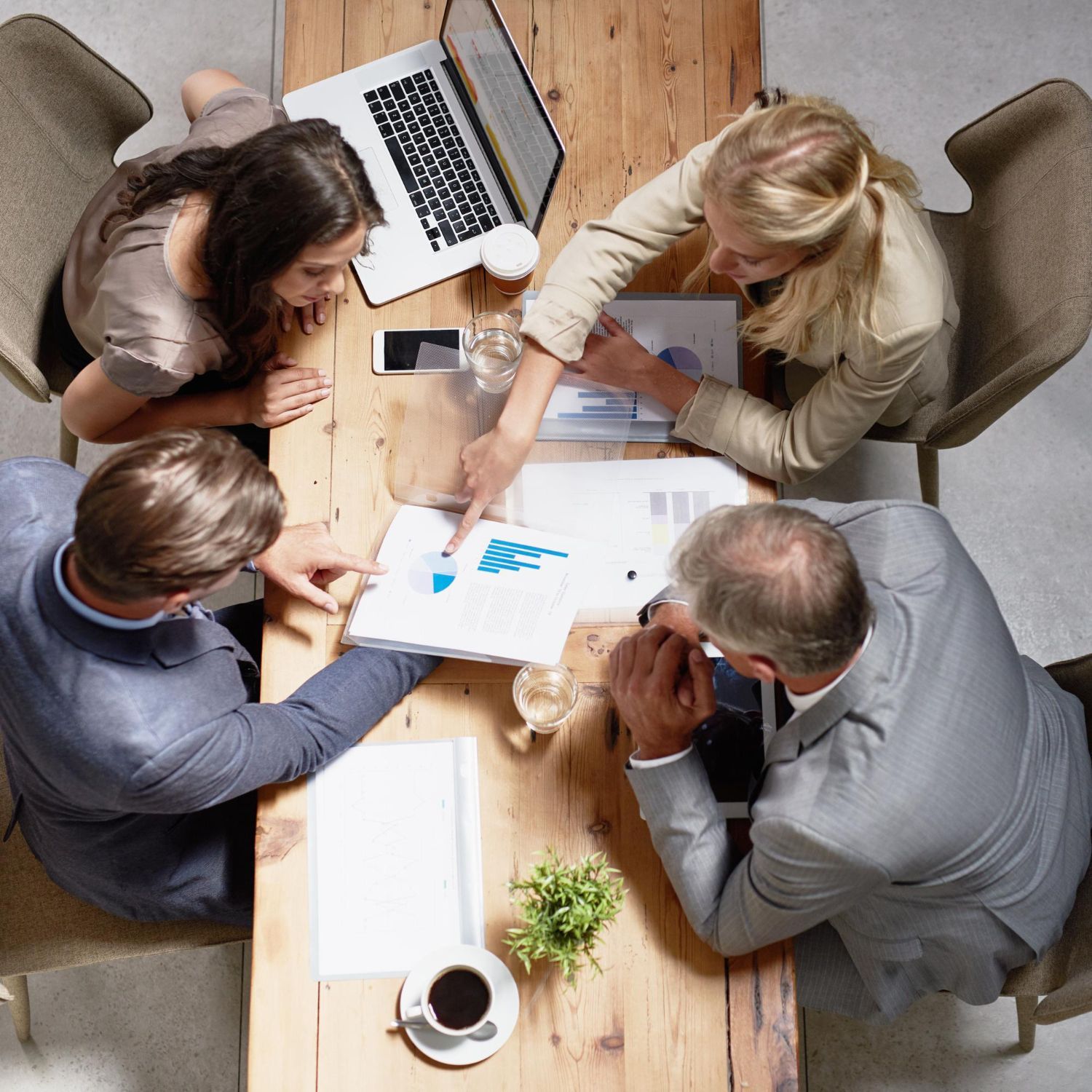 Four people at a wooden table in a high-angle view, working together on documents and a laptop during a meeting.