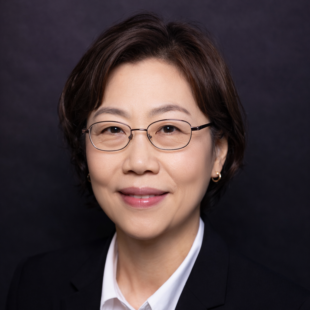 A person with a beard and short hair wears round, yellow-tinted glasses and a colorful zip-up jacket against a grey wall.