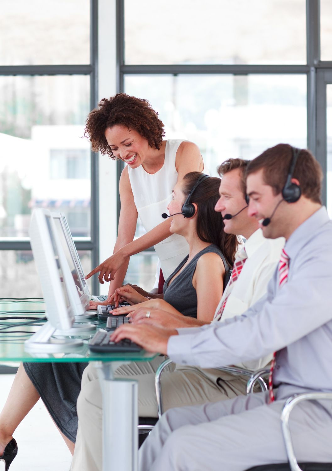 A team of customer service representatives at computers wearing headsets, with one person pointing to a screen.