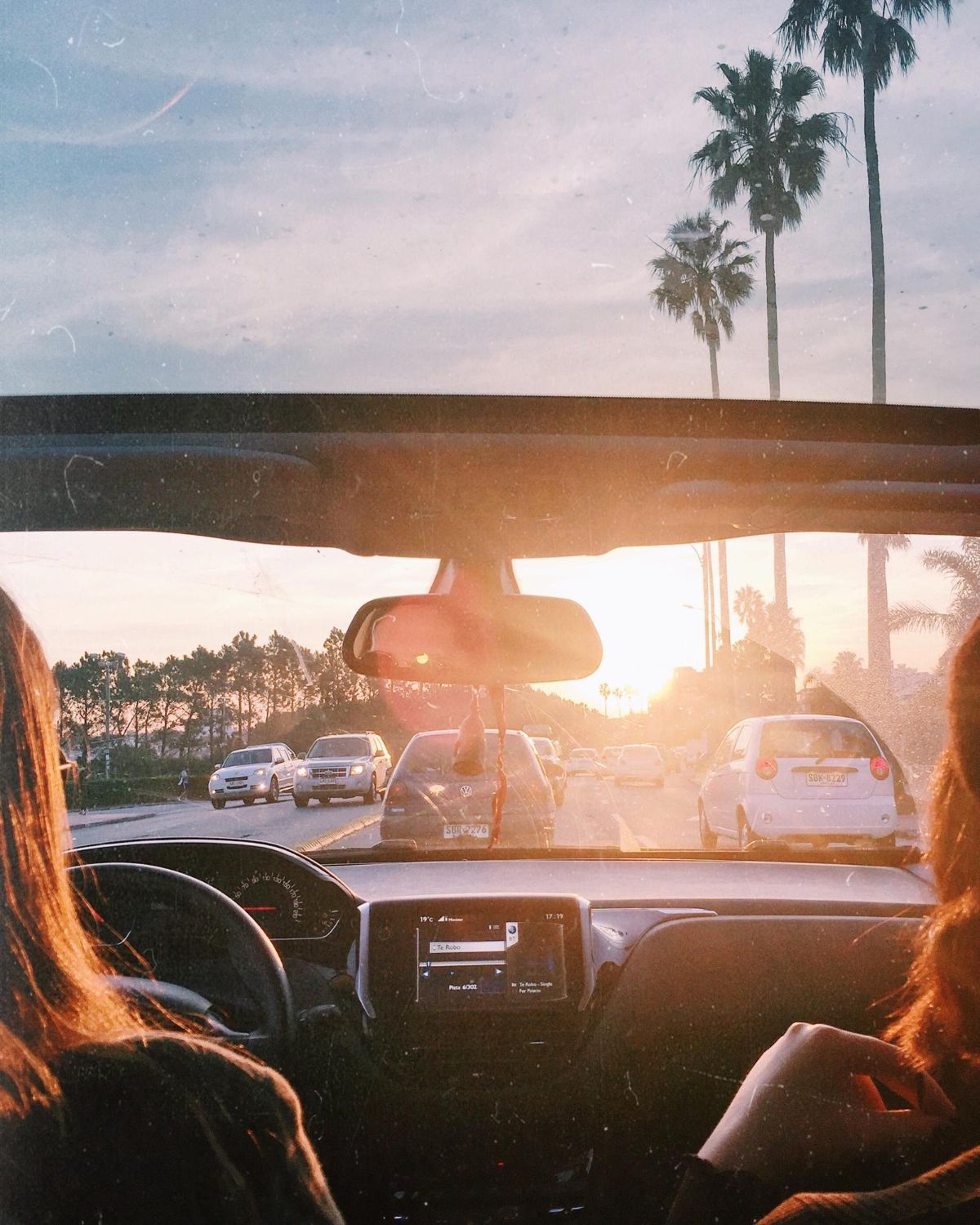 A view from inside a car driving towards a sunset, with palm trees visible under a soft light and traffic ahead.