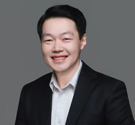 A person with freckles wears a textured yellow top and long green tassel earrings, posed against a stark white wall.