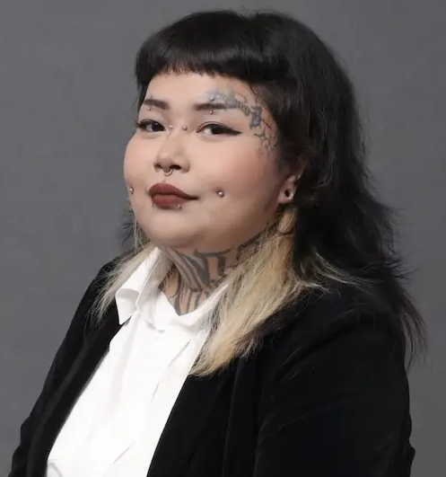 A person with freckles wears a textured yellow top and long green tassel earrings, posed against a stark white wall.