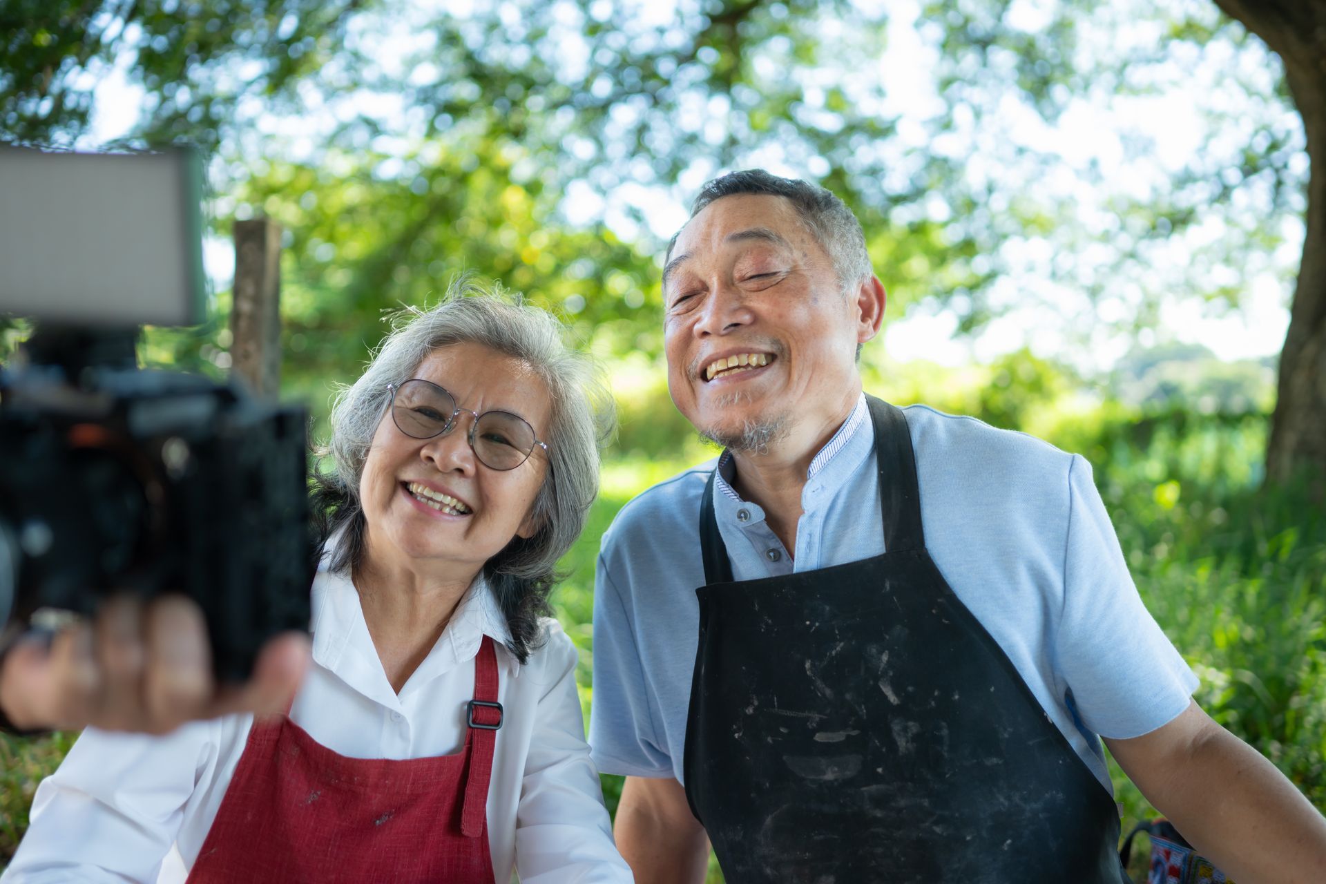 Two people wearing aprons smile while recording a video on a camera in a sunny, tree-filled outdoor setting.