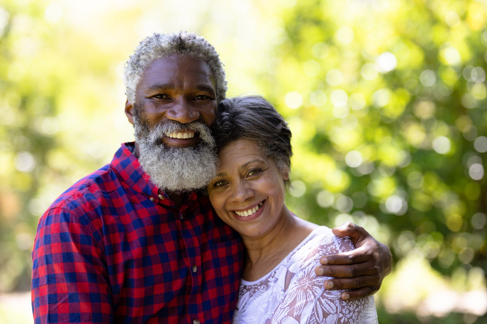 An smiling couple embracing each other outdoors against a blurred, sunlit green background.