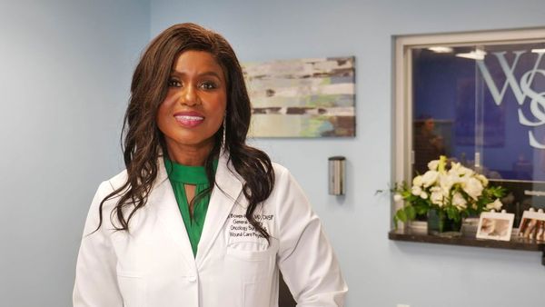 Woman in lab coat smiles in a medical office, with floral arrangement and logo in background.
