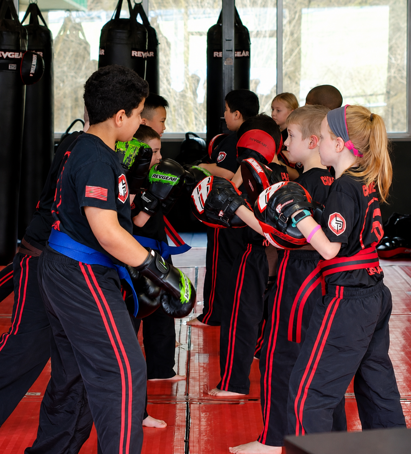Children in black-and-red martial arts uniforms training in pairs on a red mat inside a gym