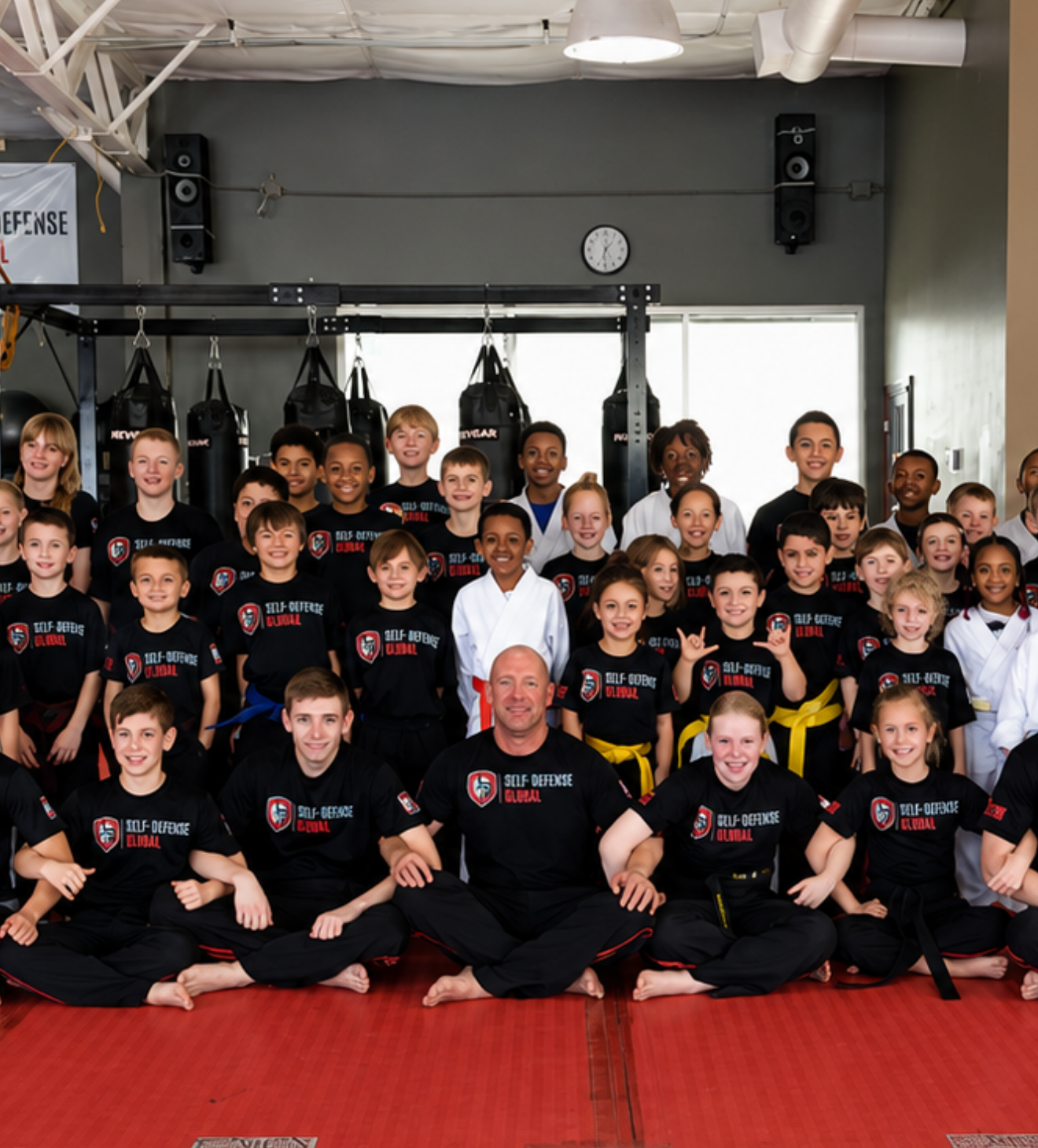 Group of martial arts students and instructors posing in a gym, wearing black uniforms on red mats.