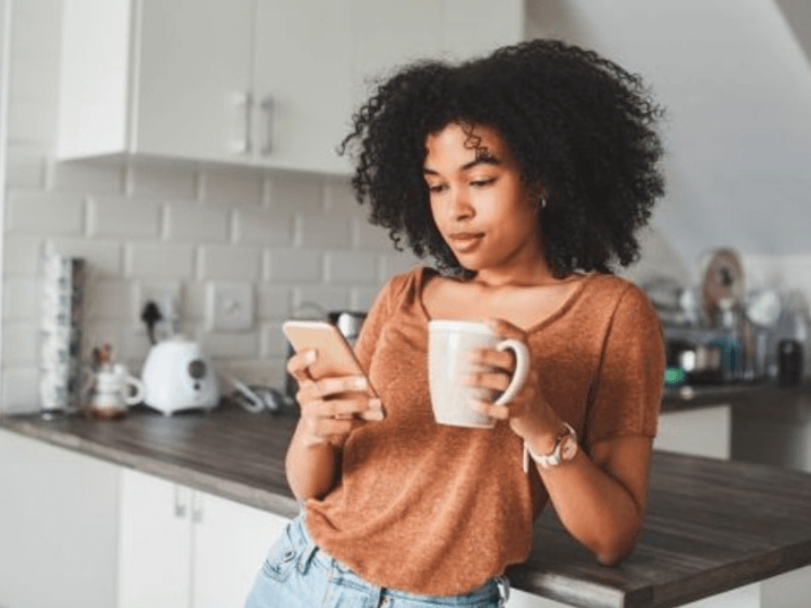 A woman is standing in a kitchen holding a cup of coffee and looking at her phone.