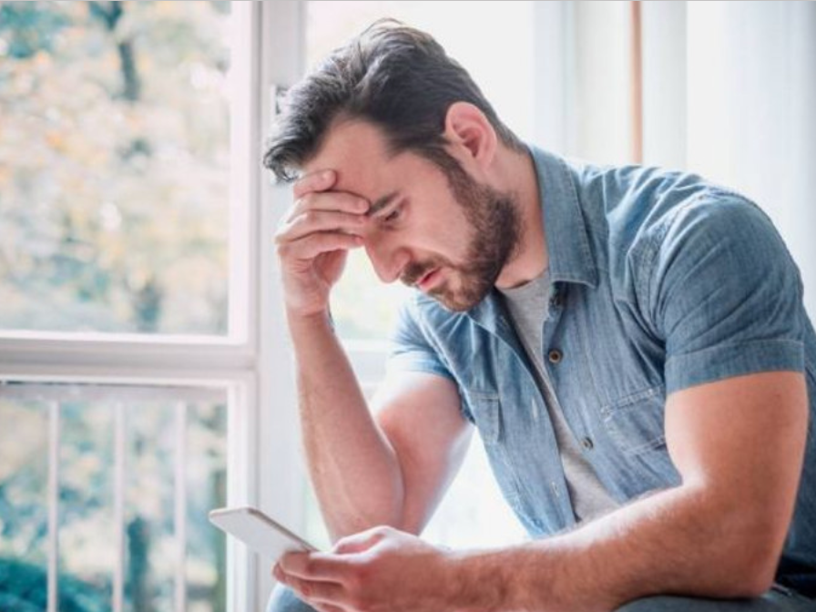 A man is sitting on a window sill looking at his cell phone.