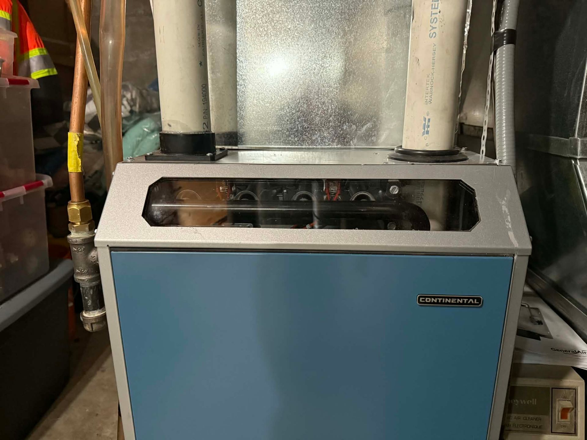 A blue and grey residential furnace sits in a basement with two white PVC pipes extending from its top.