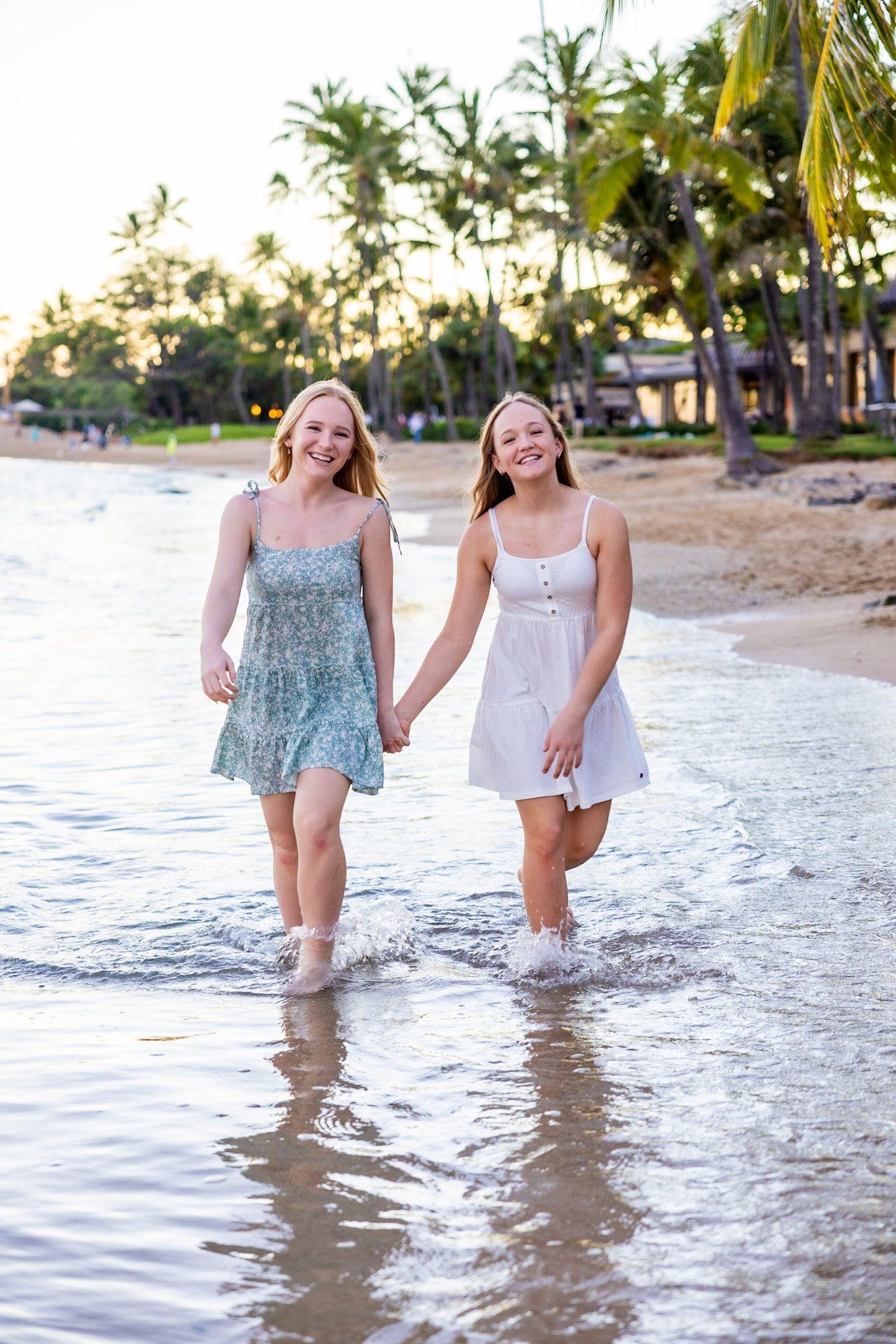 Two women are holding hands while walking through the water on the beach.