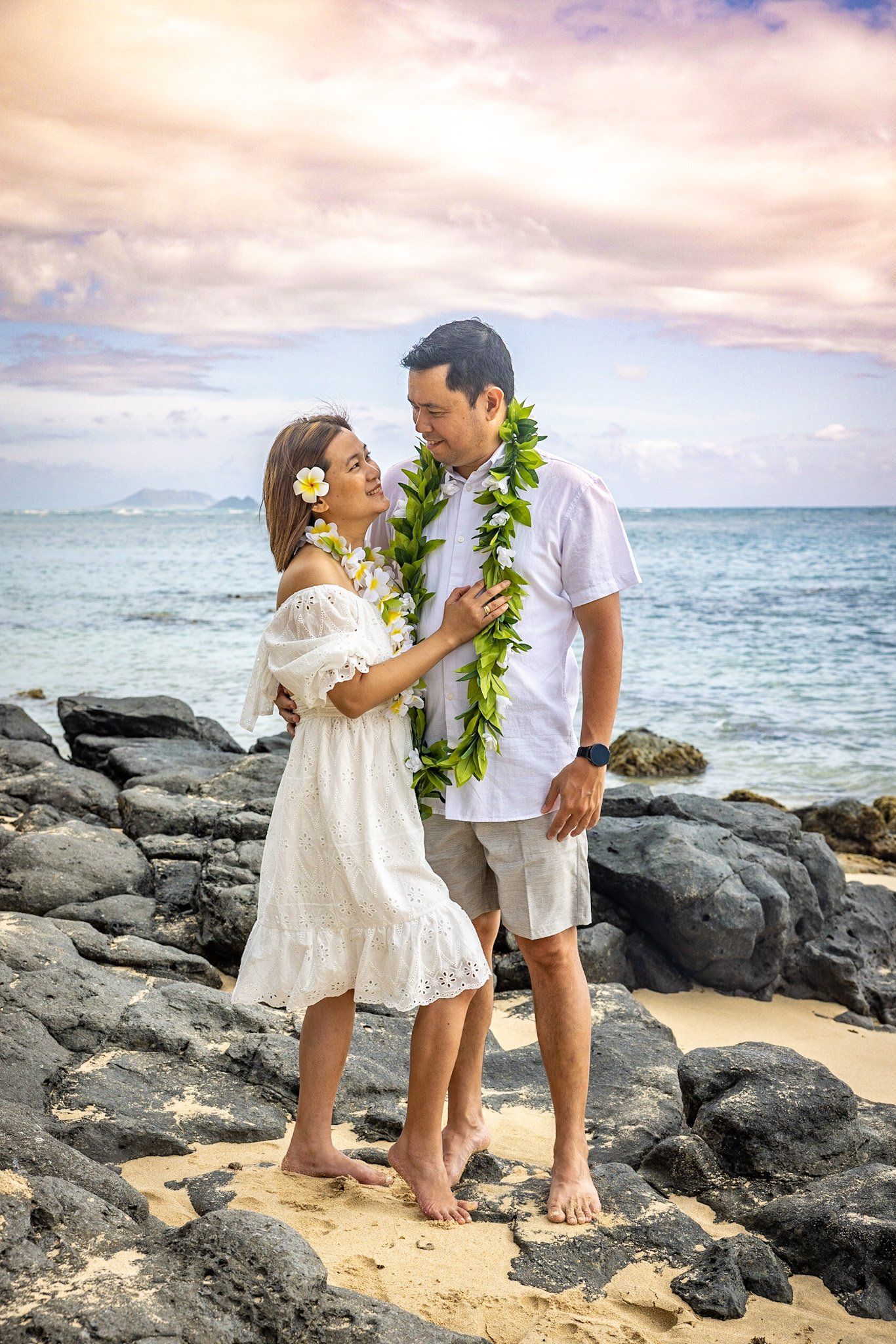 A man and a woman are standing on a rocky beach with a lei around their necks.