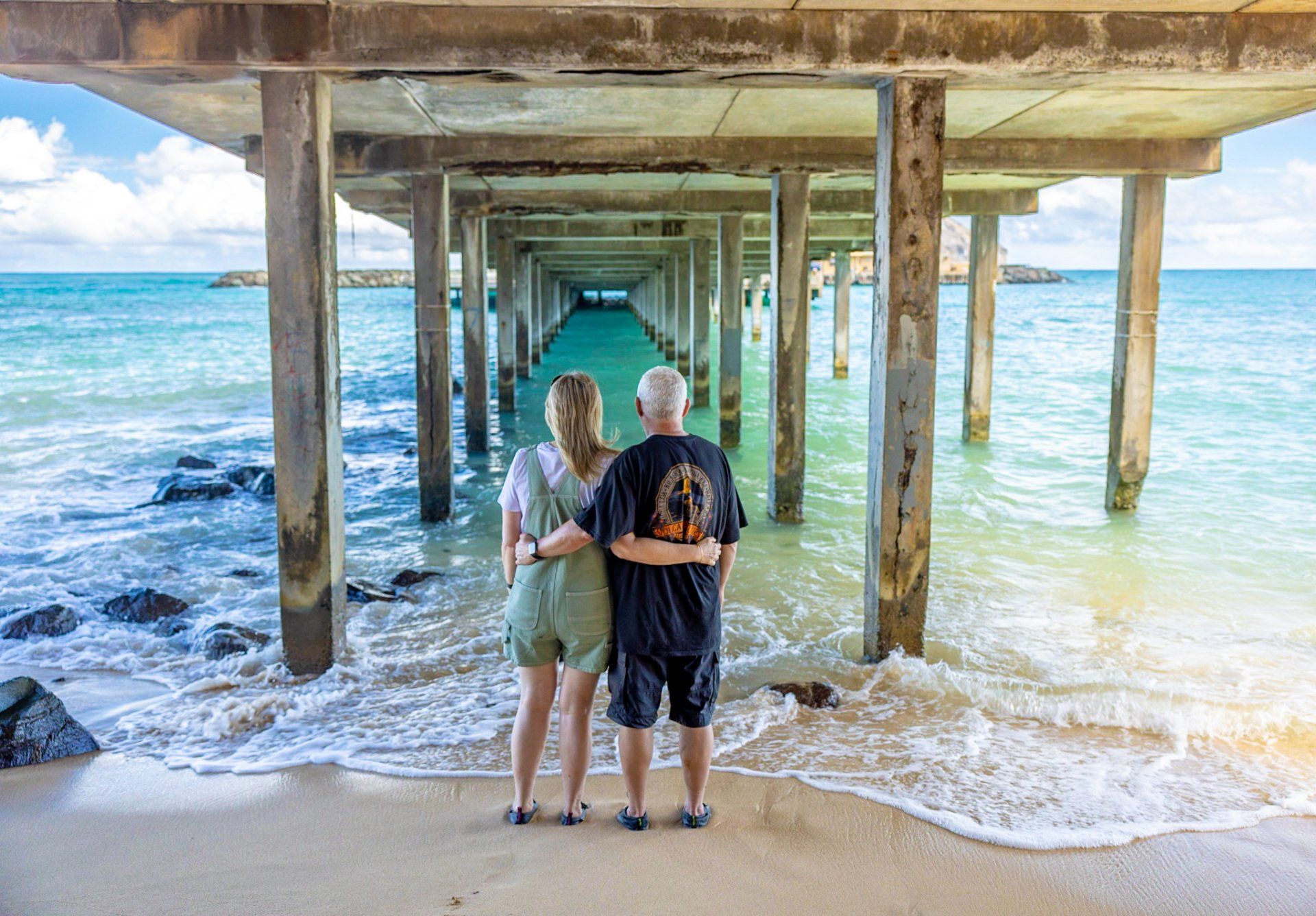 A man and a woman are standing under a pier on a beach.