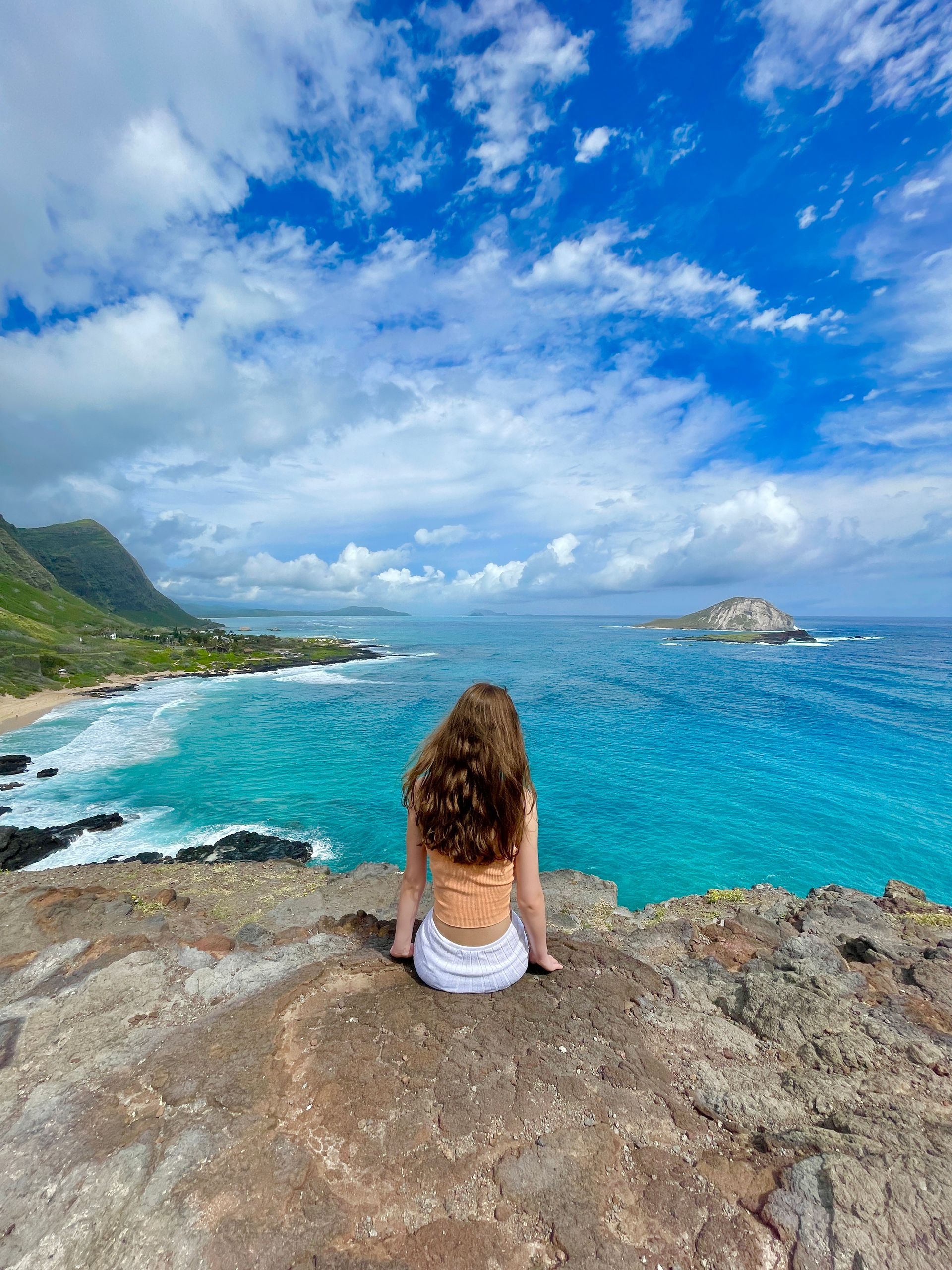 A woman is sitting on a rock overlooking the ocean.
