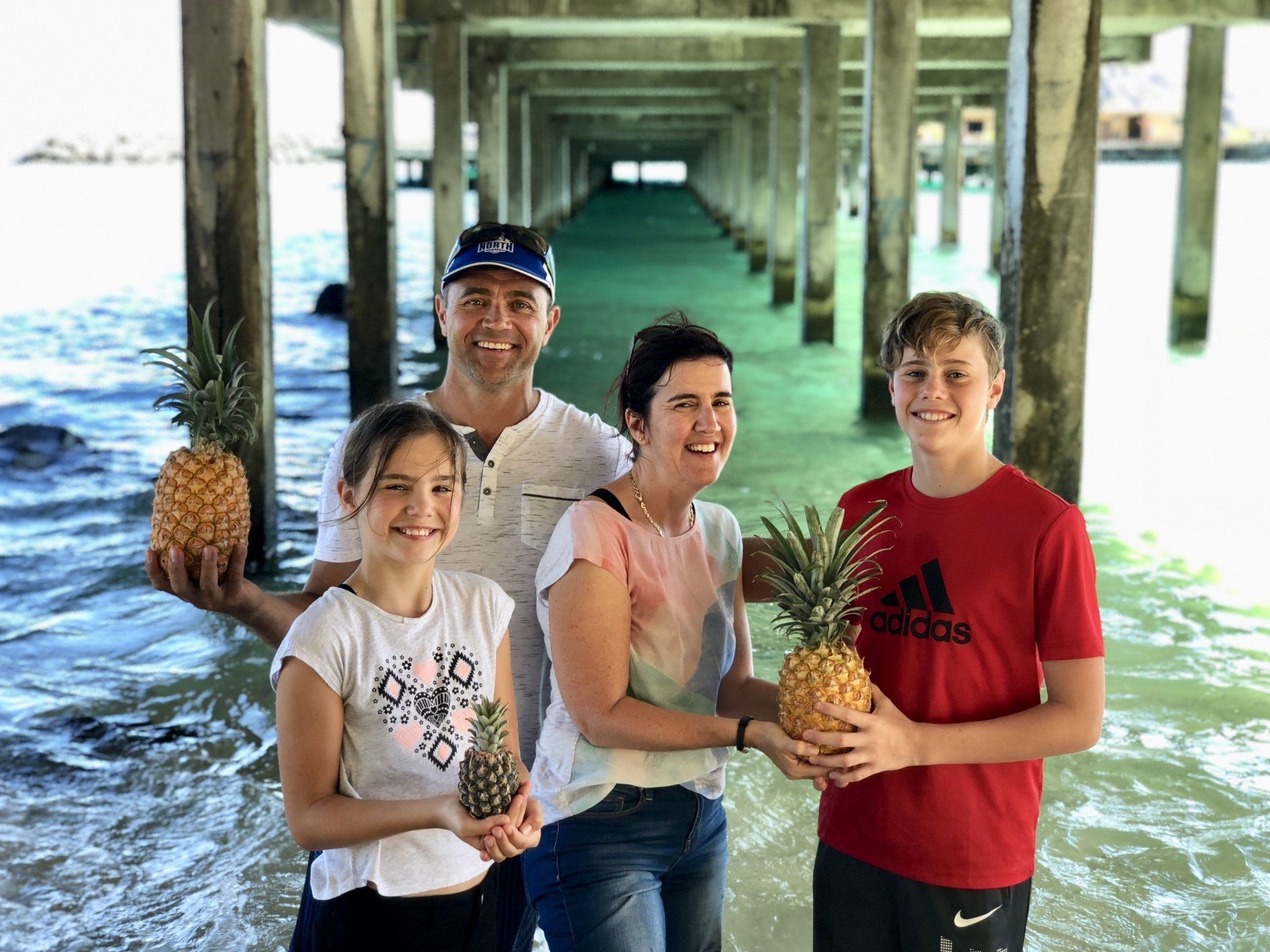 A family is standing under a pier holding pineapples.
