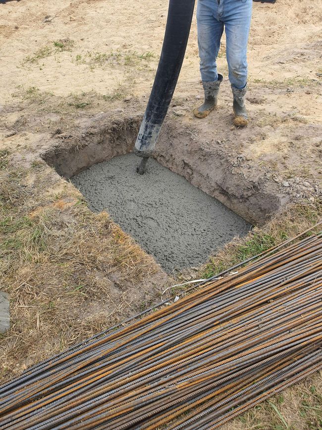 Man pours concrete into a rectangular pit, rebar laid nearby on dirt and grass.
