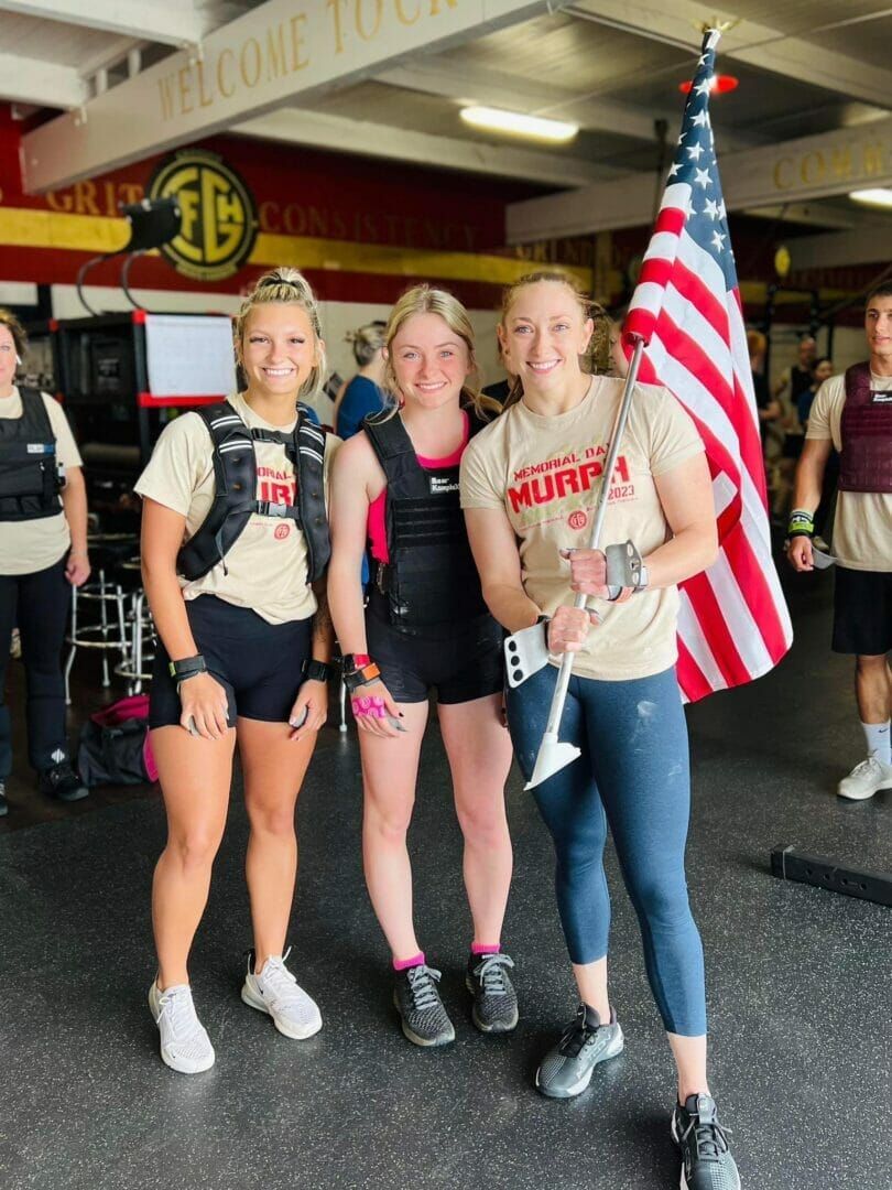 Three women are posing for a picture in front of an american flag in a gym.