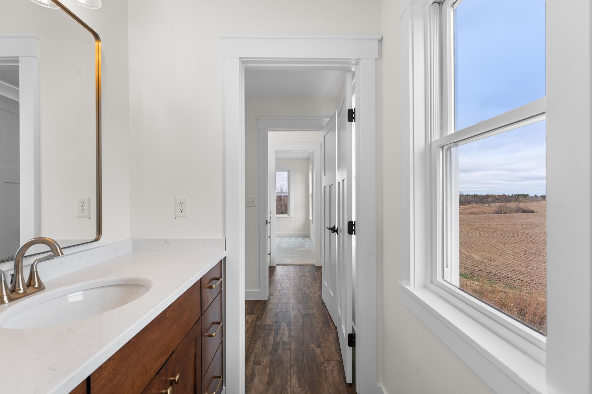 Bathroom hallway with dark wood vanity, white walls, and a window with a view of a field.