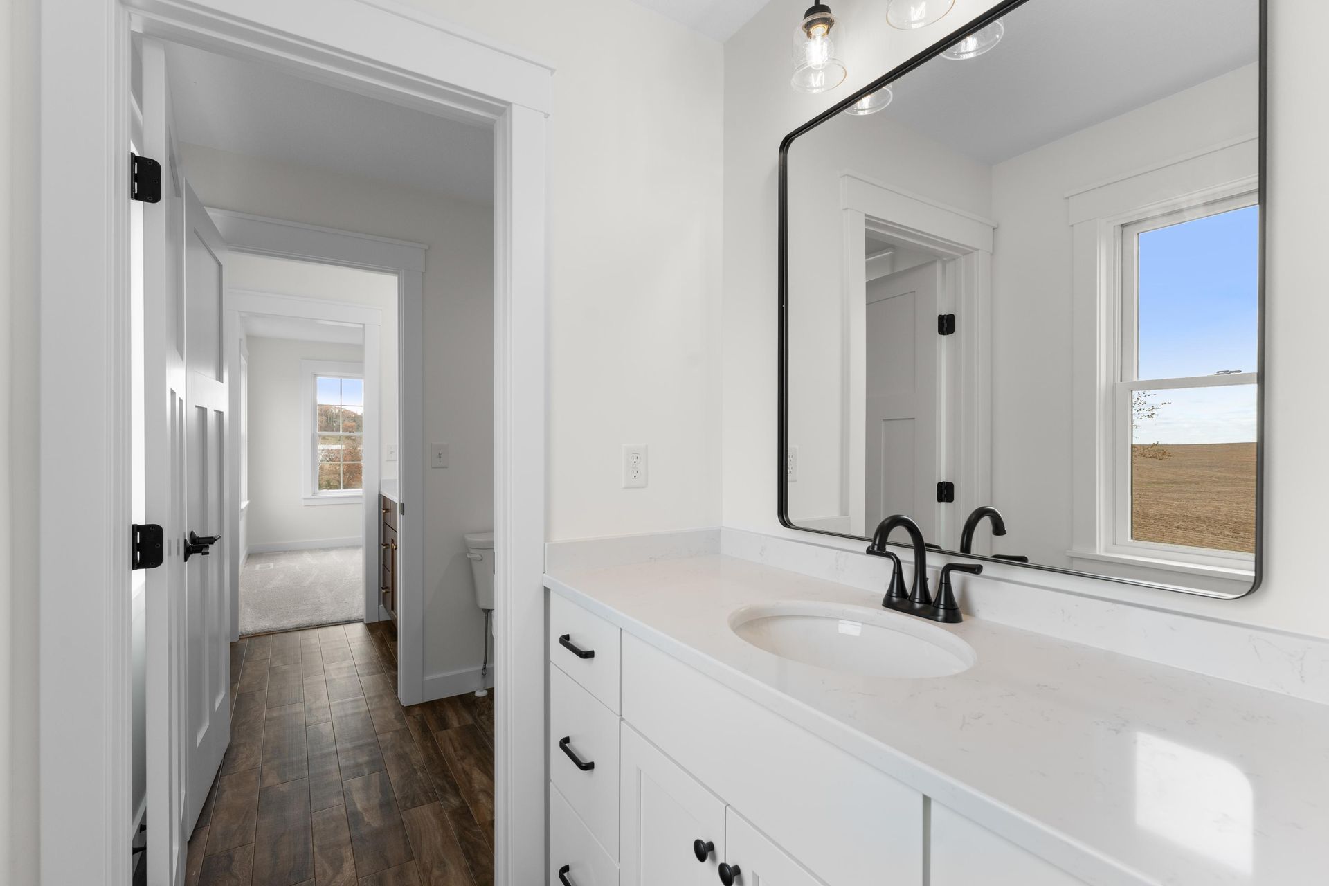 Bathroom with white cabinetry, dark fixtures, and a large mirror. A hallway leads to another room.