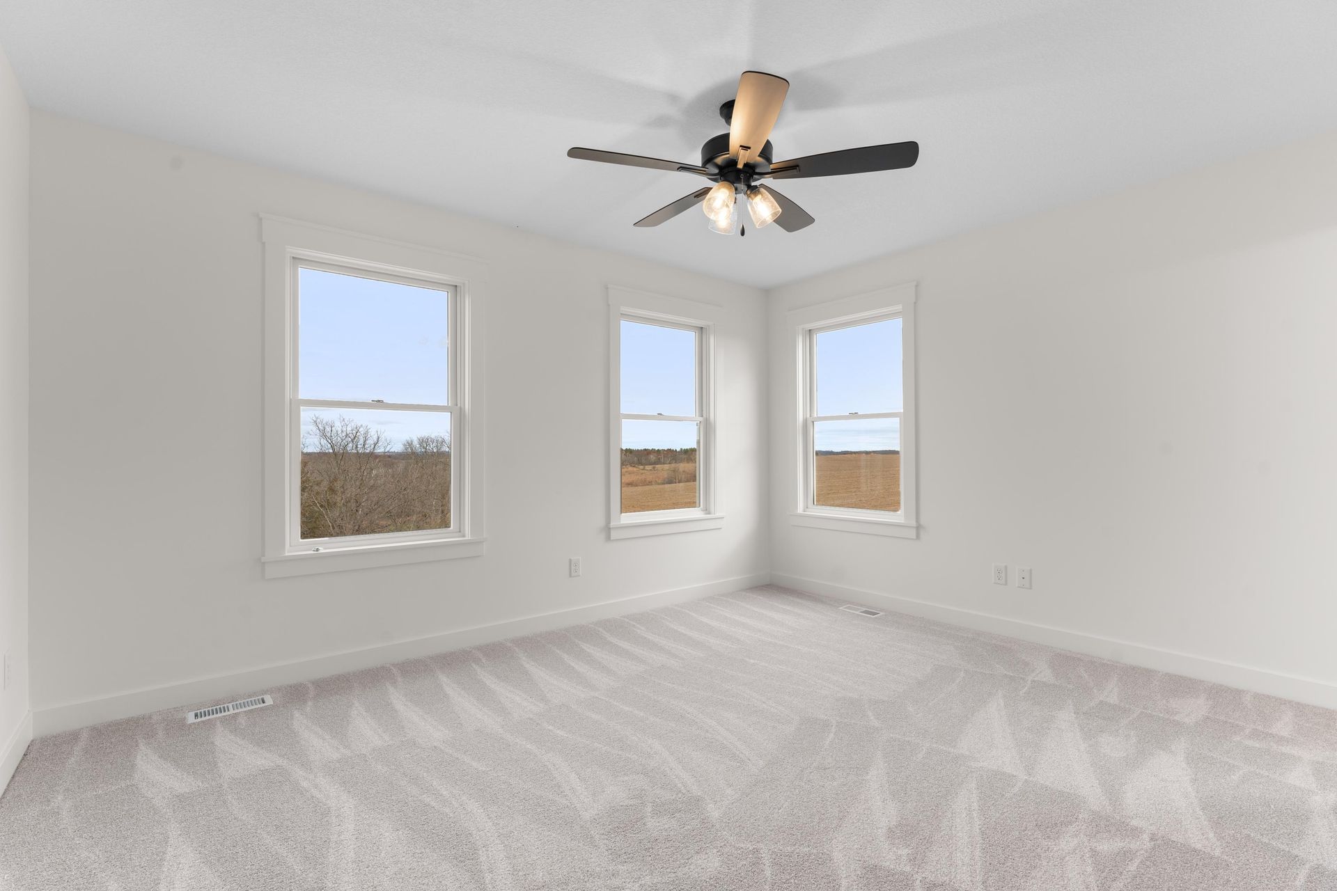Empty bedroom with light gray carpet, three windows, and a ceiling fan.