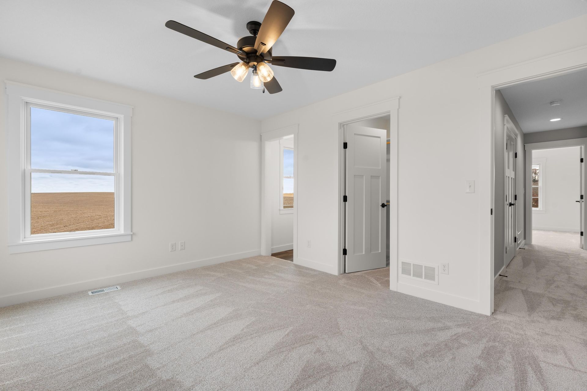 Empty bedroom with light carpet, white walls, and a ceiling fan; a window shows a view of a field.
