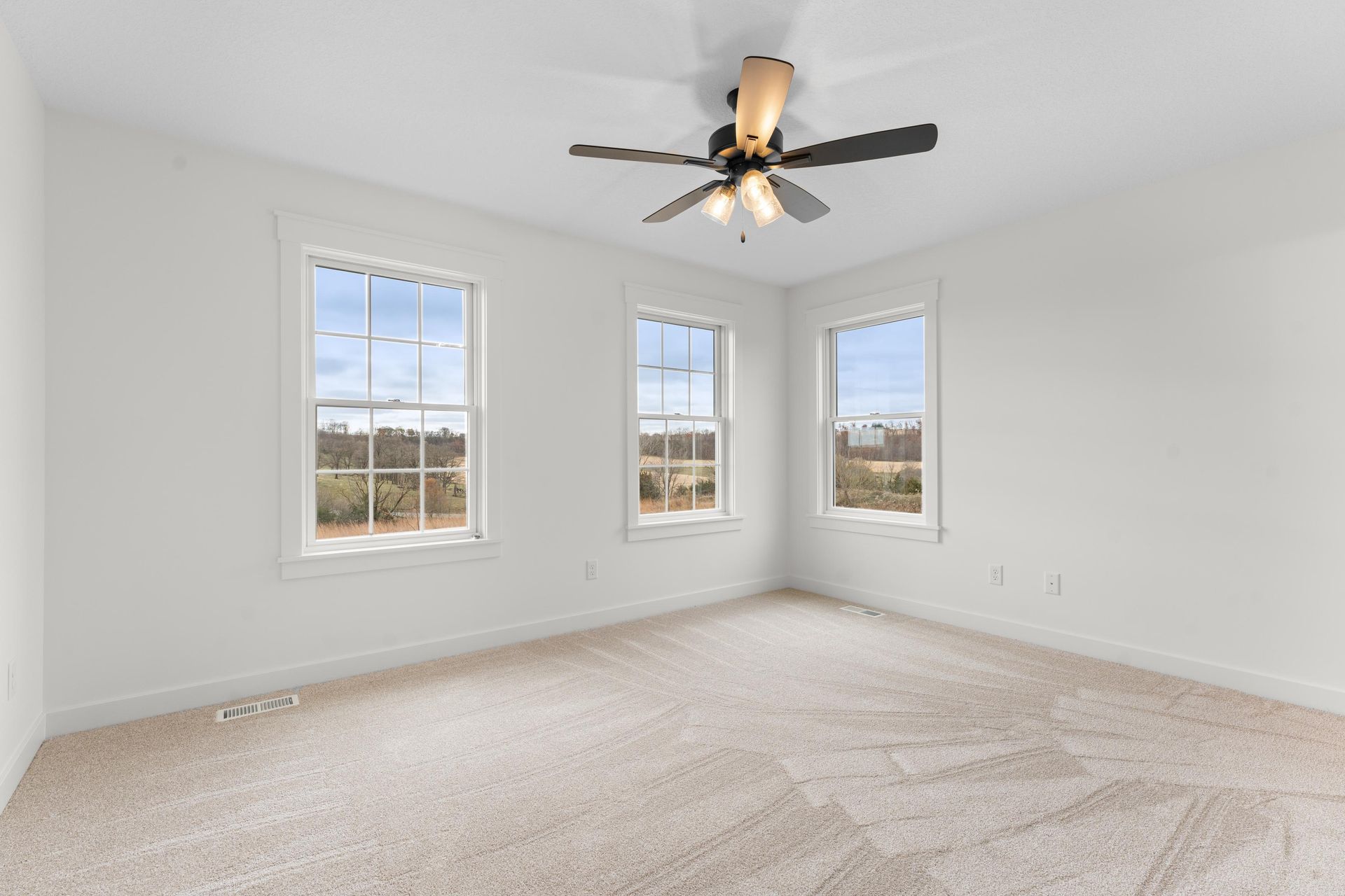 Empty white-walled room with three windows and a ceiling fan, carpeted floor.