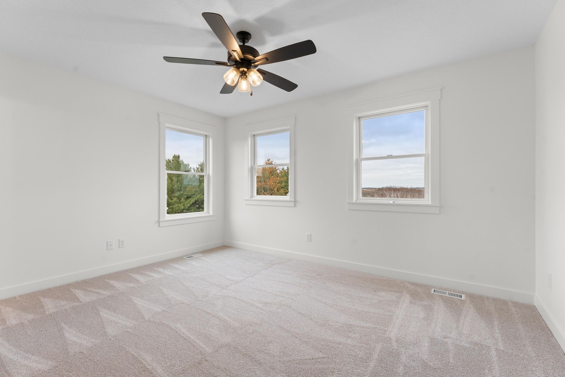 Empty bedroom with white walls, three windows, beige carpet, and a ceiling fan.