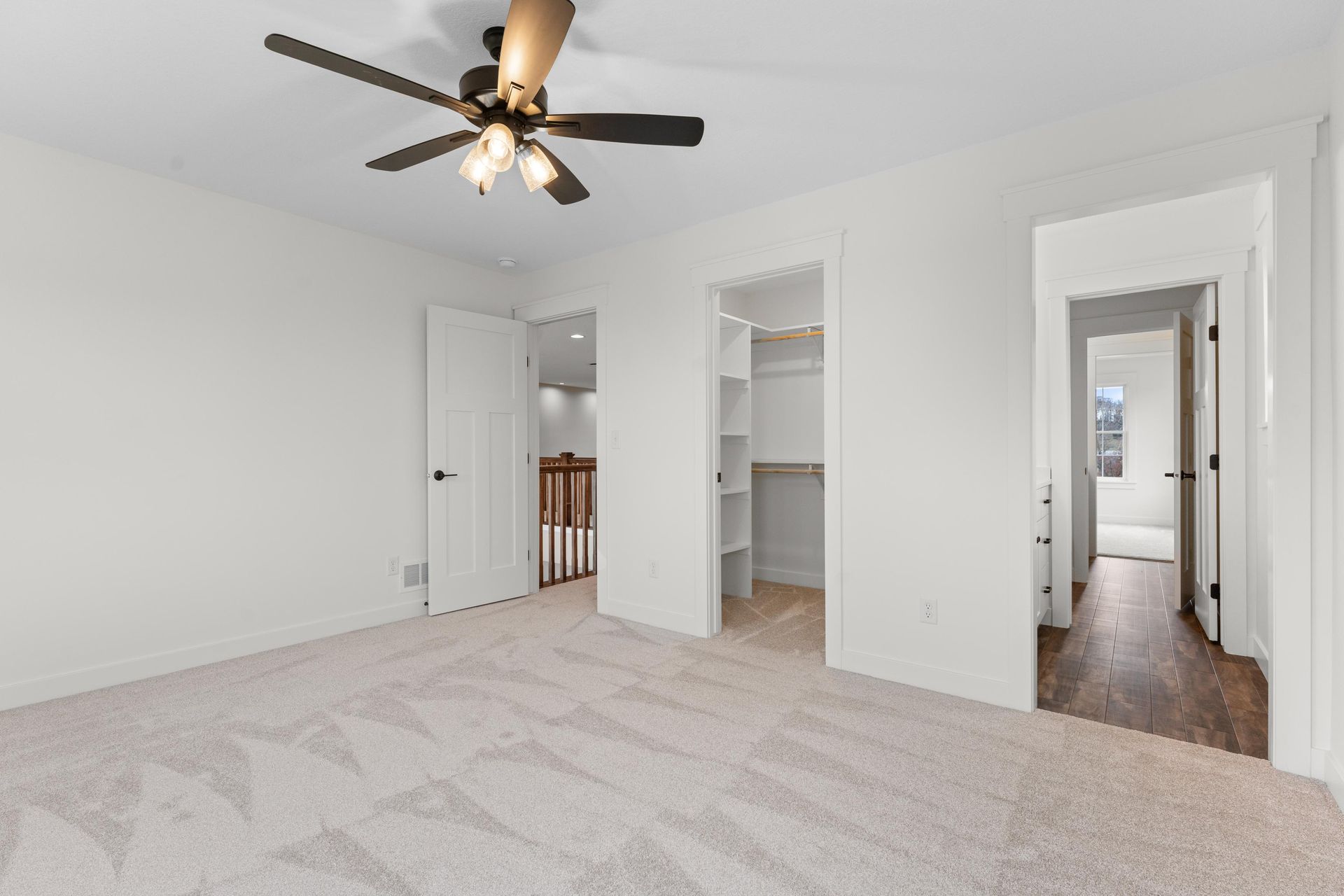 Empty white bedroom with carpet, ceiling fan, doors to closet, hall, and stairs.