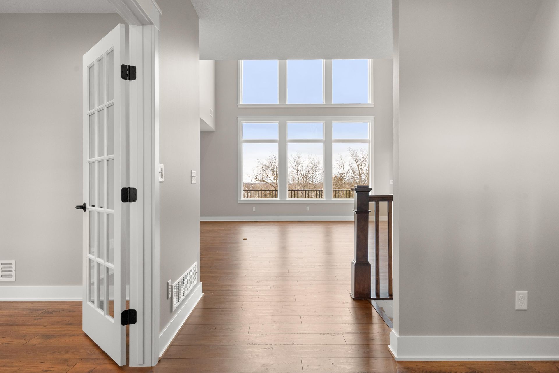 Interior hallway with wooden floor, white trim, and a door opening to a bright living room with large windows.