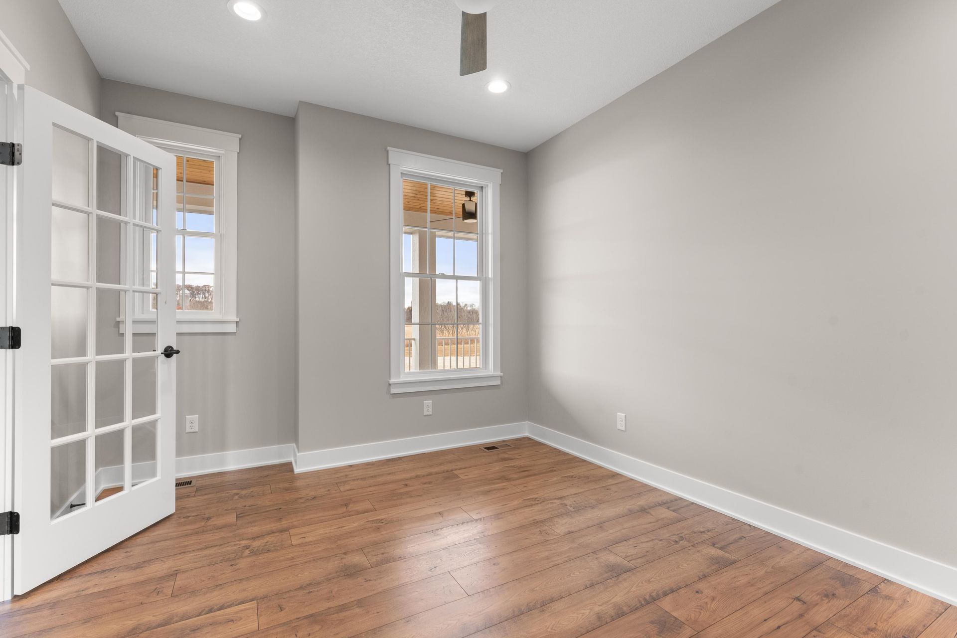 Empty room with hardwood floors, two windows, and French doors. Light gray walls, white trim.