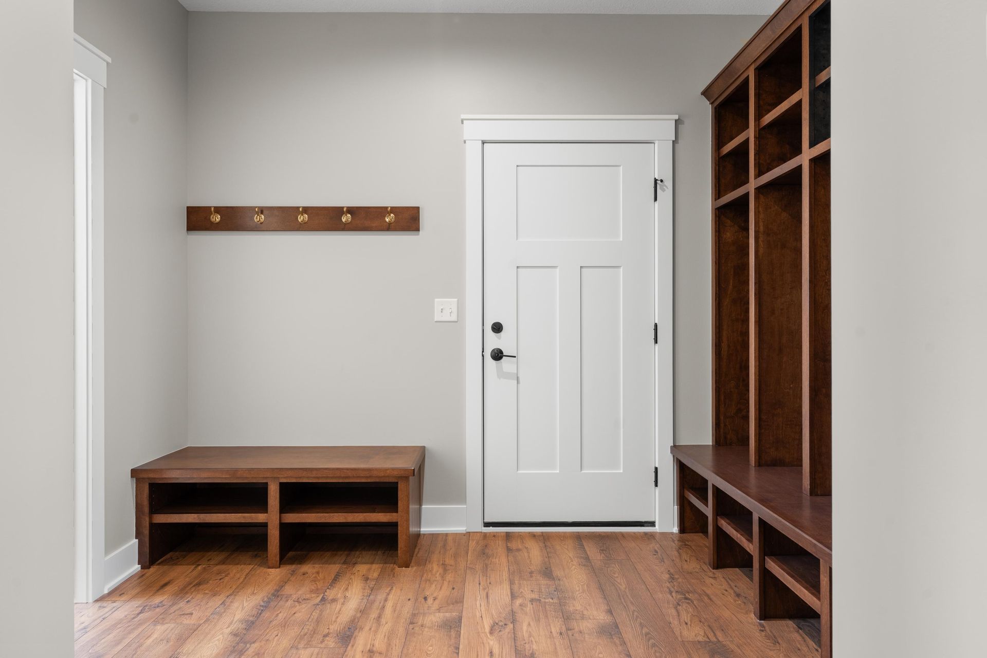Mudroom with wood bench, coat rack, door, and built-in cubbies. Light gray walls, dark wood trim and flooring.