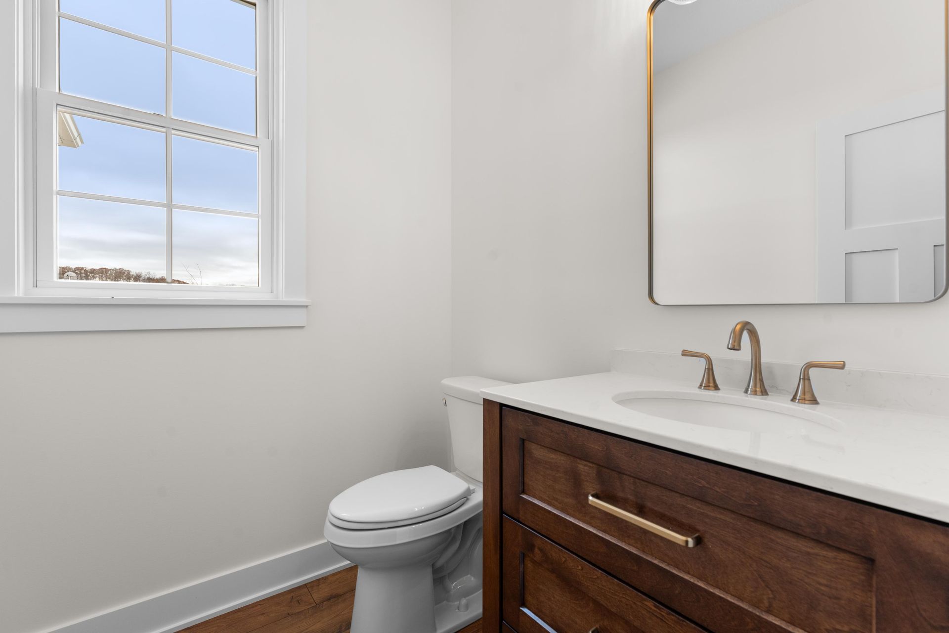 Bathroom with wooden vanity, white countertop, gold fixtures, toilet, and window.