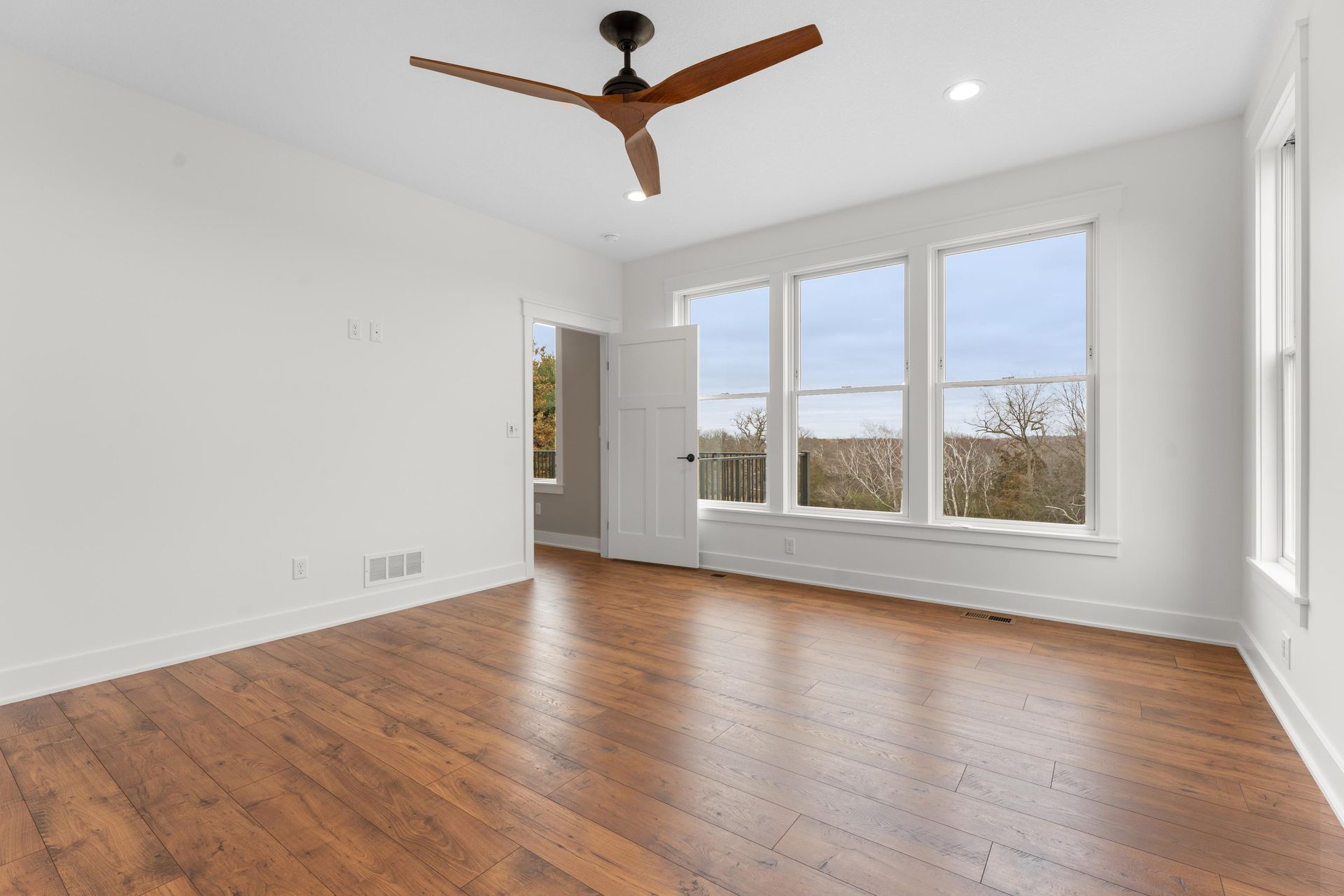 Empty, bright room with hardwood floors, large windows, ceiling fan, and open door to an outside view.