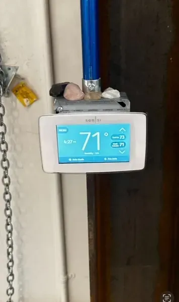 A thermostat with a blue screen is hanging on a pipe.