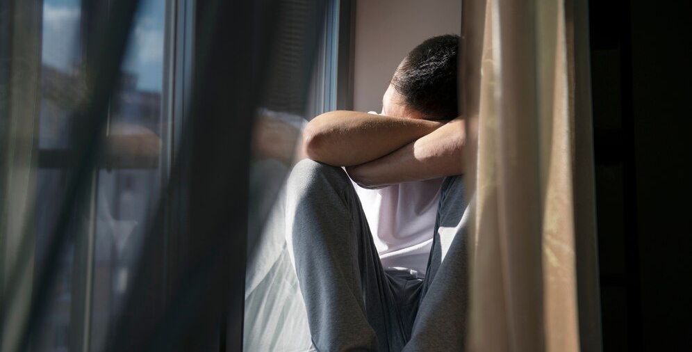 A man is sitting on a window sill with his head in his hands.