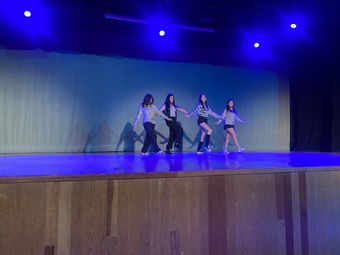 Cuatro personas tomadas de la mano, bailando en un escenario bajo focos azules, con paneles de madera en primer plano.