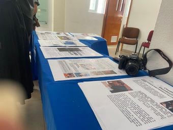 Una mesa cubierta con un mantel azul muestra páginas impresas y una cámara. Vista interior.