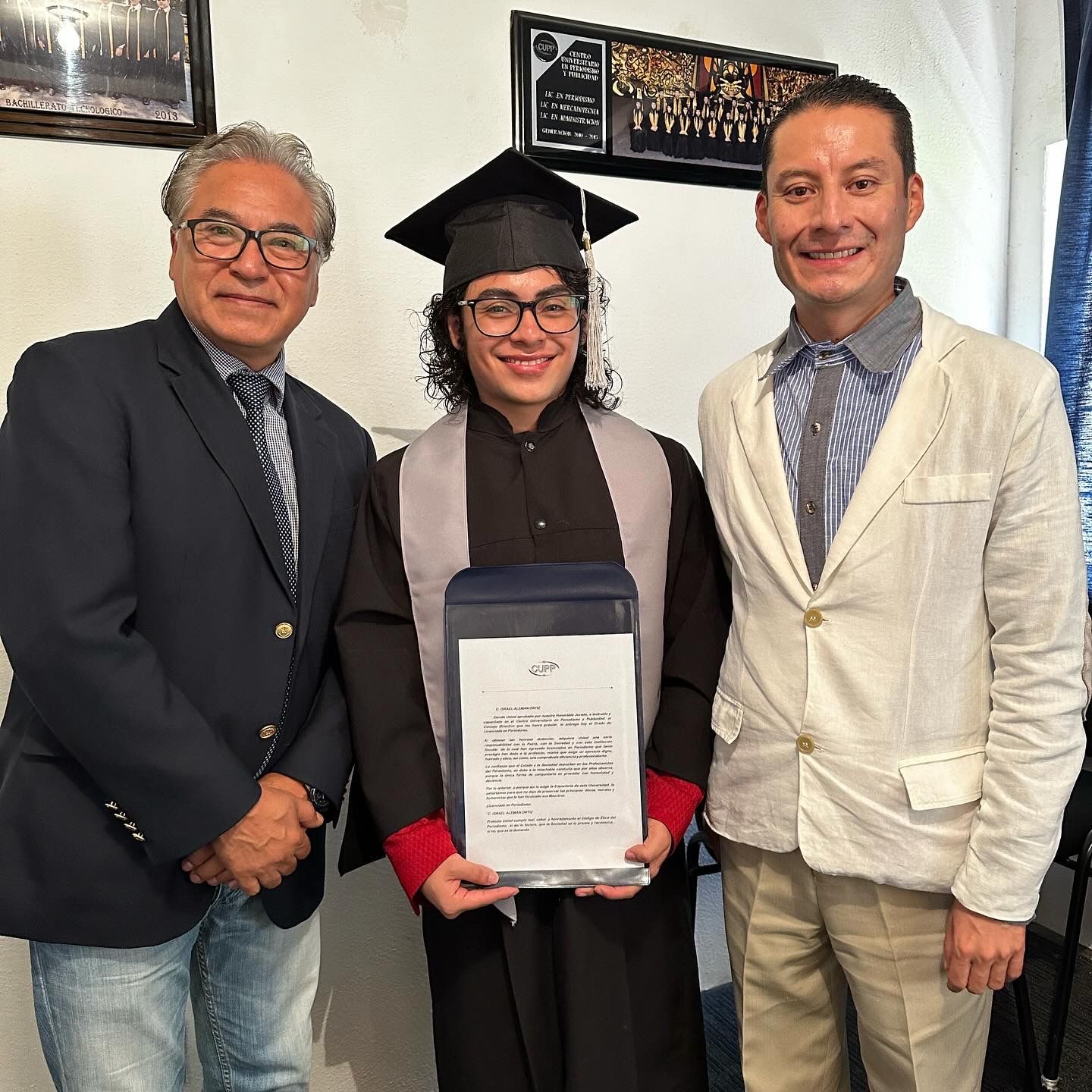 Tres personas: un graduado con toga y birrete sosteniendo un certificado, flanqueado por dos hombres, en el interior.