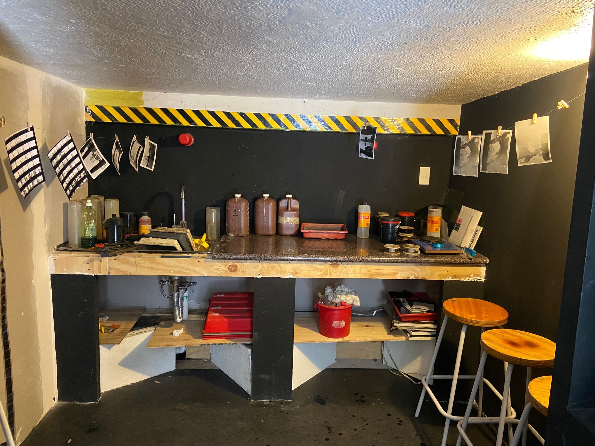 Taller con banco de trabajo, herramientas y taburetes. Pared negra con franja amarilla de peligro.